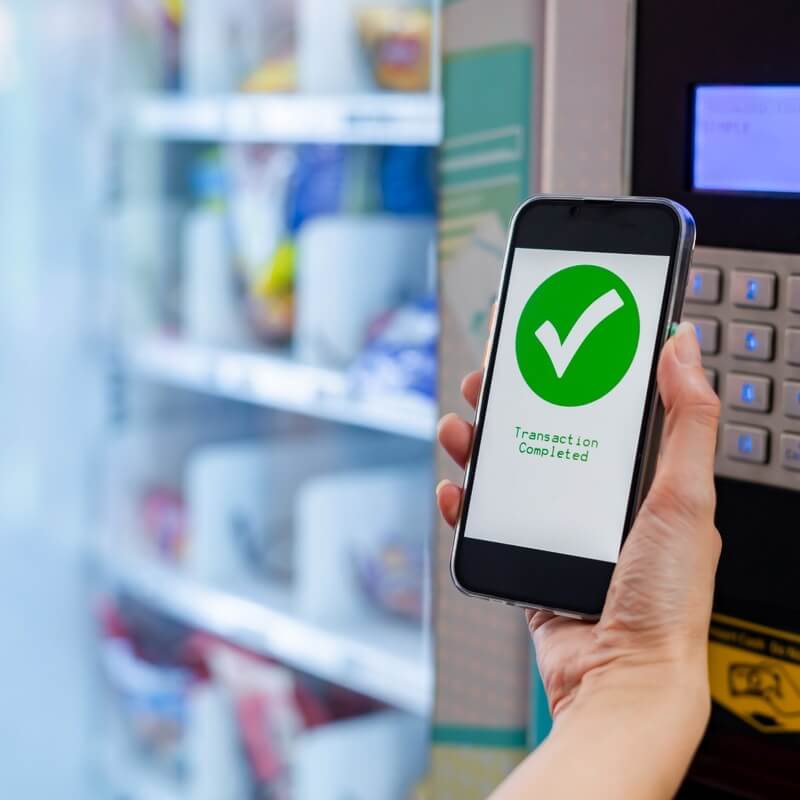 In front of a vending machine, a person holds a phone where it reads "transaction completed".