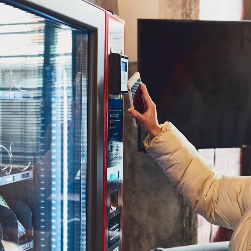 In front of a vending machine, a person holds a phone next to the payment terminal.