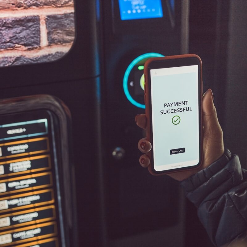 A woman holds a smartphone where it reads "payment successful" in front of a vending machine.