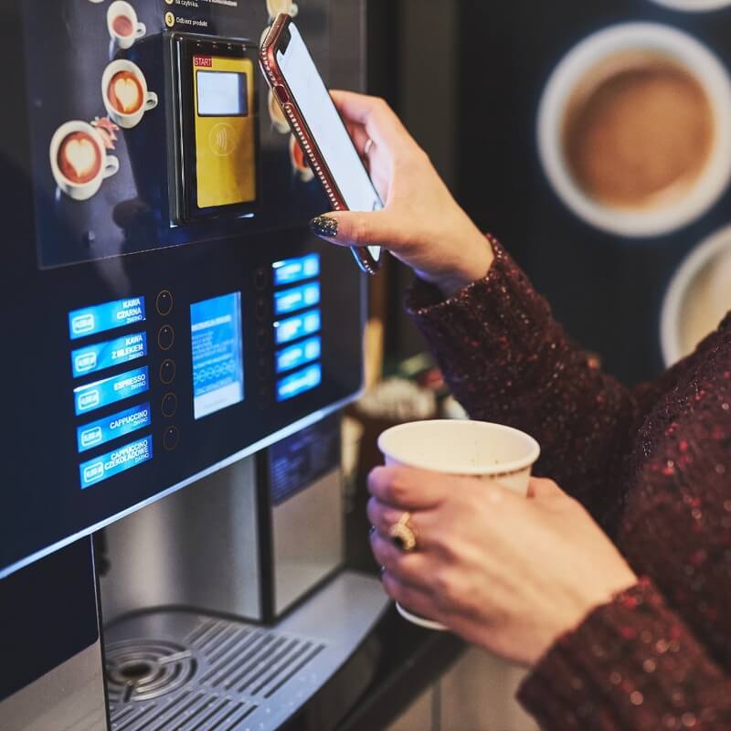 A woman holds her smartphone close to the payment terminal of a vending machine that dispenses drinks. In her other hand, she holds a cup of coffee.